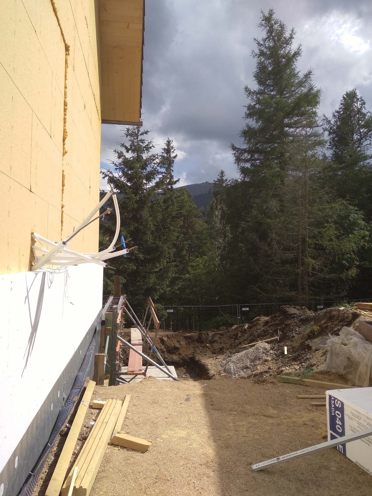 Ocieplanie ściany budynku styropianem z widocznymi elementami konstrukcyjnymi, rurami i deskami na placu budowy w tle z drzewami i pochmurnym niebem.