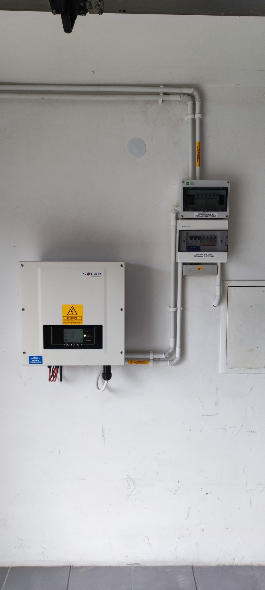 Biały inwerter Sofar Solar zamontowany na białej ścianie obok rozdzielnicy elektrycznej z białymi rurkami instalacyjnymi.