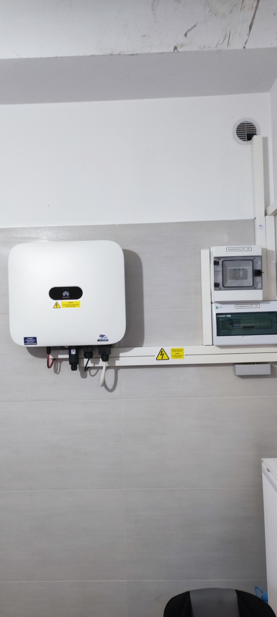 Biały falownik Huawei zamontowany na ścianie z szarych płytek, obok rozdzielnica PV-AC i PV-DC w białej obudowie, widoczne przewody i żółta naklejka ostrzegawcza.