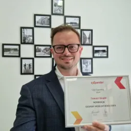 Uśmiechnięty mężczyzna w okularach i marynarce prezentuje nominację do nagrody Ekspert Mobilny Roku 2023 firmy Expander na tle ściany z czarno-białymi zdjęciami w ramkach.