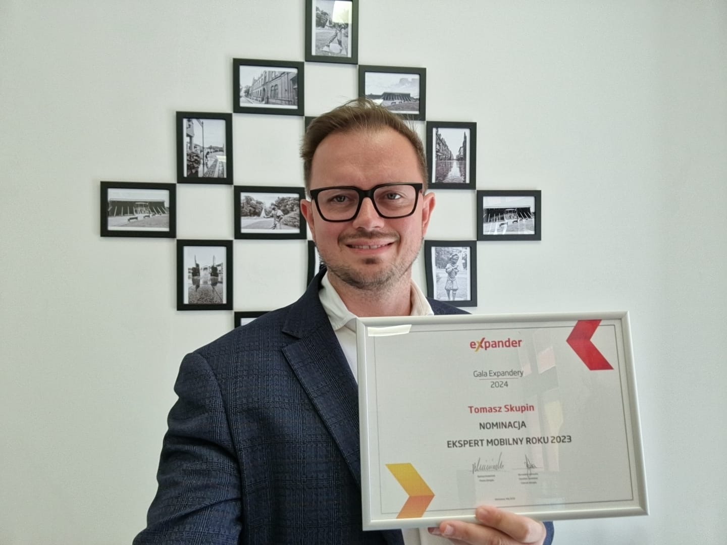 Uśmiechnięty mężczyzna w okularach i marynarce prezentuje nominację do nagrody Ekspert Mobilny Roku 2023 firmy Expander na tle ściany z czarno-białymi zdjęciami w ramkach.