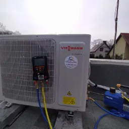 Biała jednostka zewnętrzna klimatyzacji Viessmann DC Inverter, zamontowana na betonowych wspornikach na płaskim dachu, podłączona do niebieskiej pompy próżniowej VALUE i miernika ciśnienia Testo...