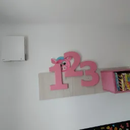 Biała jednostka rekuperacyjna na ścianie w pokoju dziecięcym, obok dekoracji z cyframi i półki z książkami.