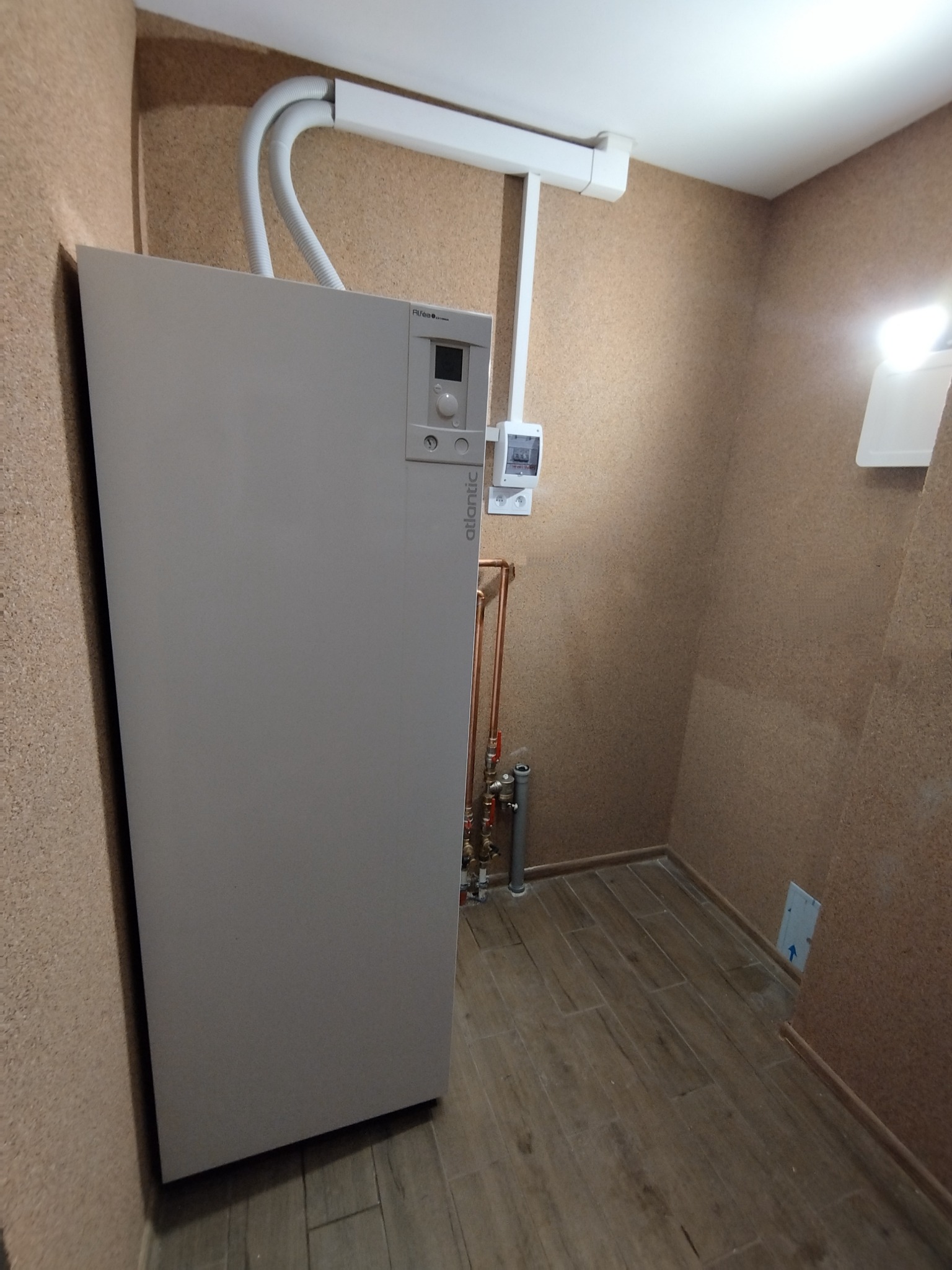 Szara pompa ciepła Atlantic z widocznymi rurami miedzianymi i białymi przewodami w pomieszczeniu wykończonym korkiem i drewnopodobną podłogą.