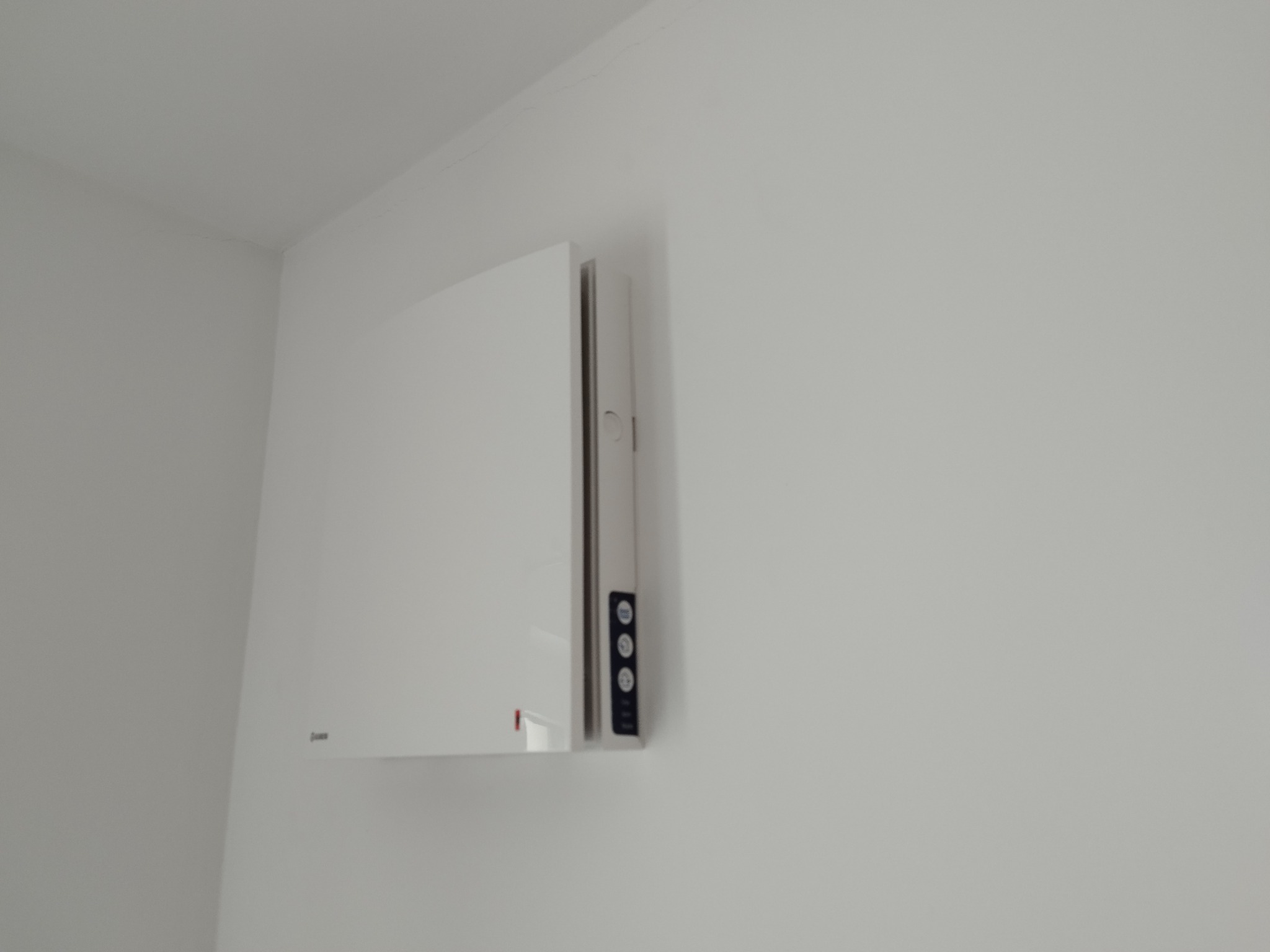 Nowoczesny, biały rekuperator ścienny z widocznym panelem sterowania zamontowany w rogu pomieszczenia o białych ścianach.