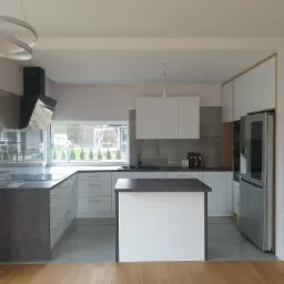 Nowoczesna kuchnia z wyspą, białe szafki i szary blat, okno z widokiem na zieleń, oświetlenie punktowe i dekoracyjny okrągły żyrandol.