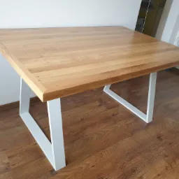 Kwadratowy stół z jasnym blatem drewnianym i białymi, metalowymi nogami w kształcie litery V, ustawiony na podłodze imitującej drewno.