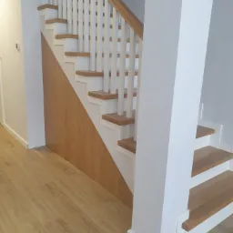 Nowoczesne schody drewniane z białymi balustradami i drewnianymi stopniami, z zabudową pod schodami w kolorze jasnego drewna, widok z dołu.