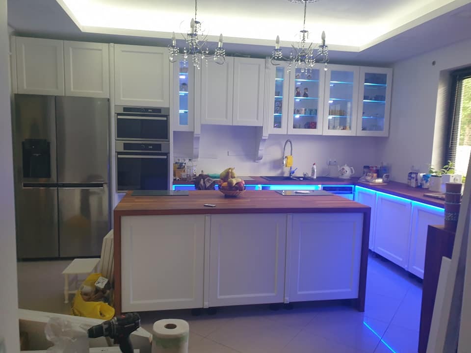 Jasna kuchnia z wyspą i oświetleniem LED, widoczne narzędzia i materiały budowlane sugerują montaż w toku.