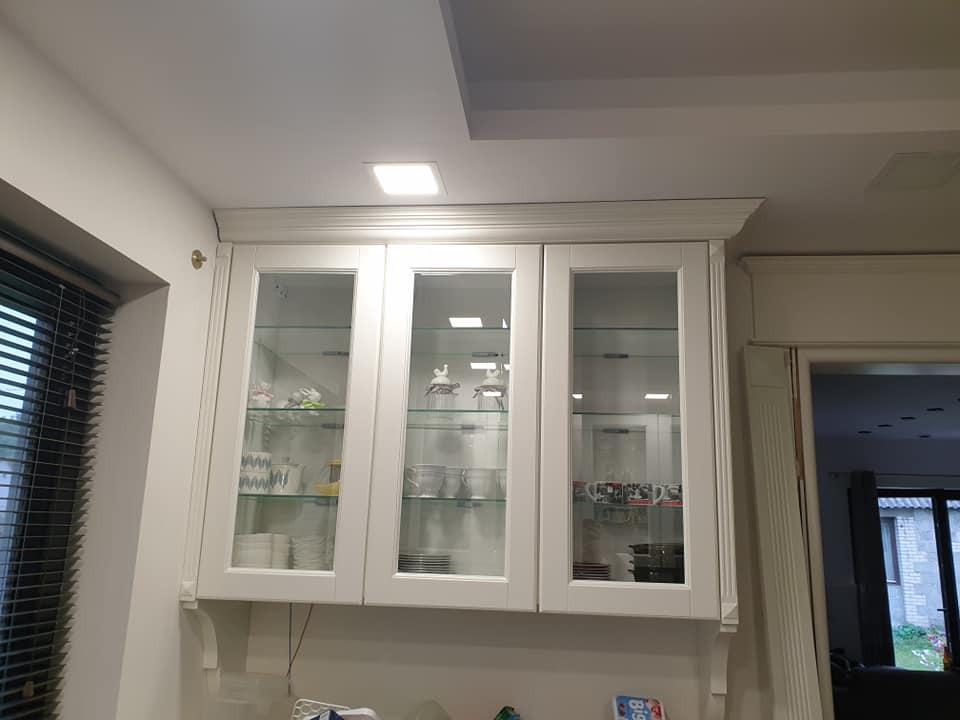 Biała wisząca szafka kuchenna z przeszklonymi drzwiami, klasyczny styl, widoczne naczynia i dekoracje wewnątrz, oświetlenie sufitowe.