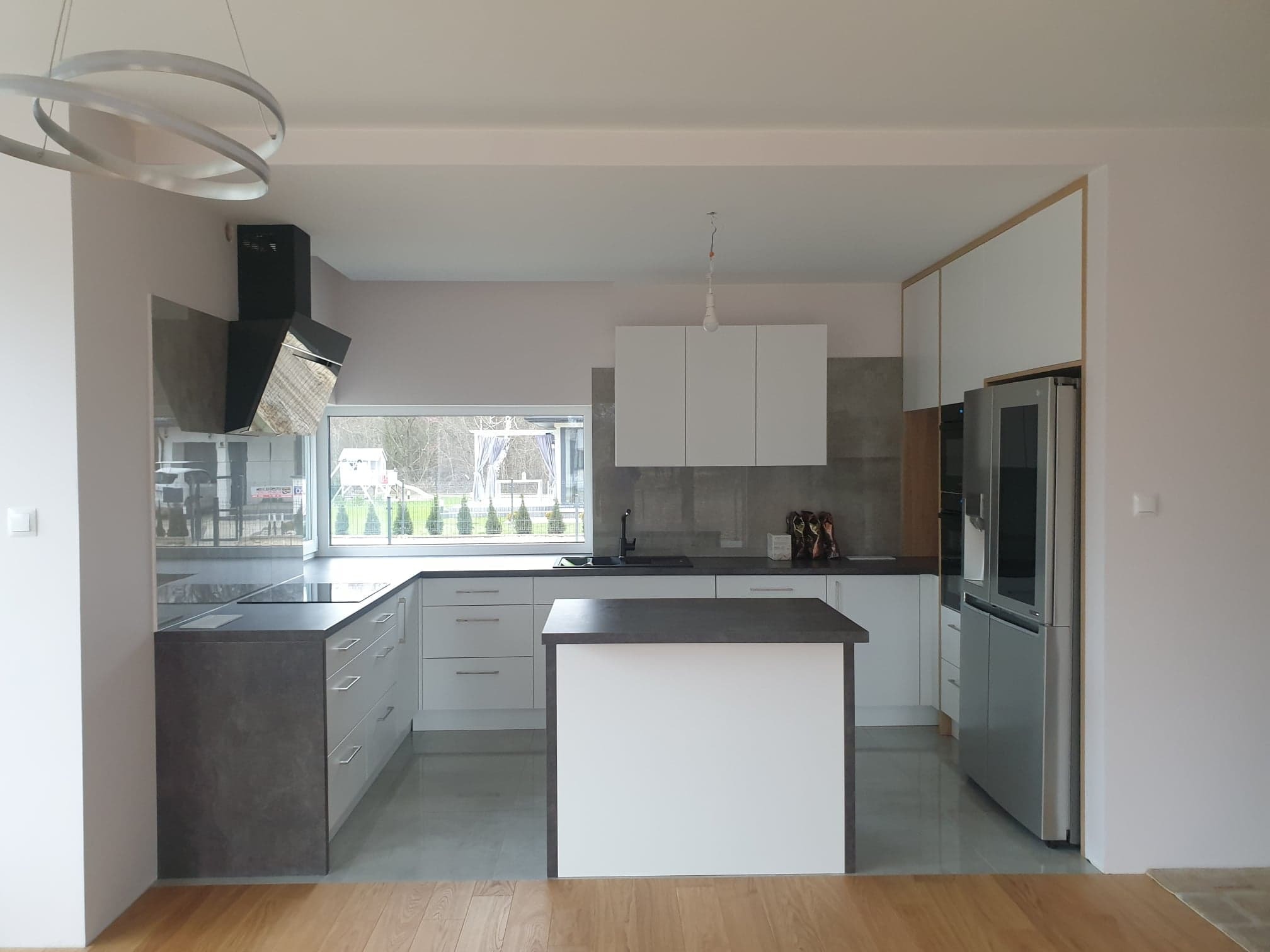 Nowoczesna kuchnia z wyspą, białe szafki i szary blat, okno z widokiem na zieleń, oświetlenie punktowe i dekoracyjny okrągły żyrandol.