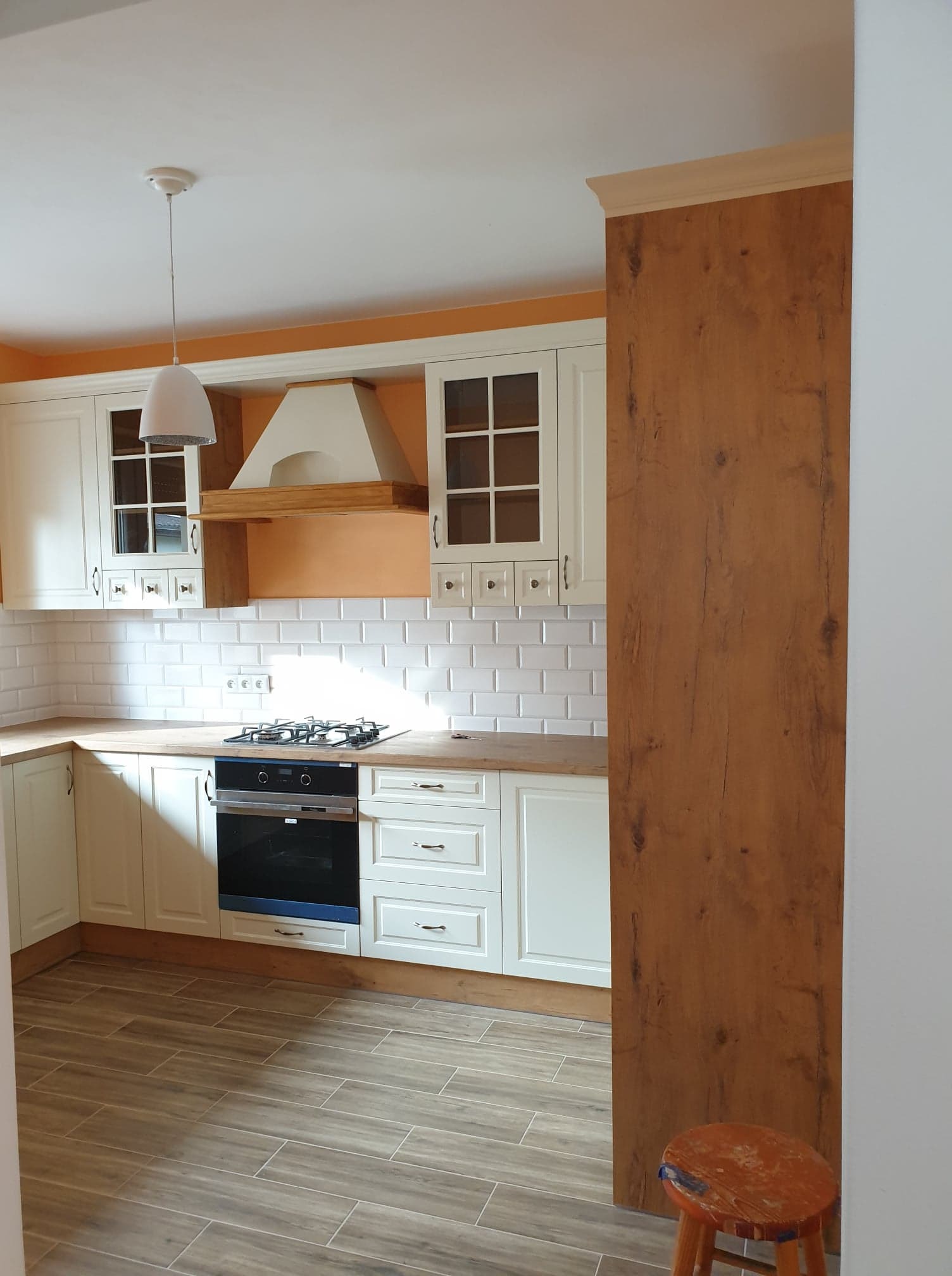 Jasna kuchnia z białymi szafkami, drewnianym blatem, płytkami metro i brązową ścianą boczną, widoczny fragment stołka.