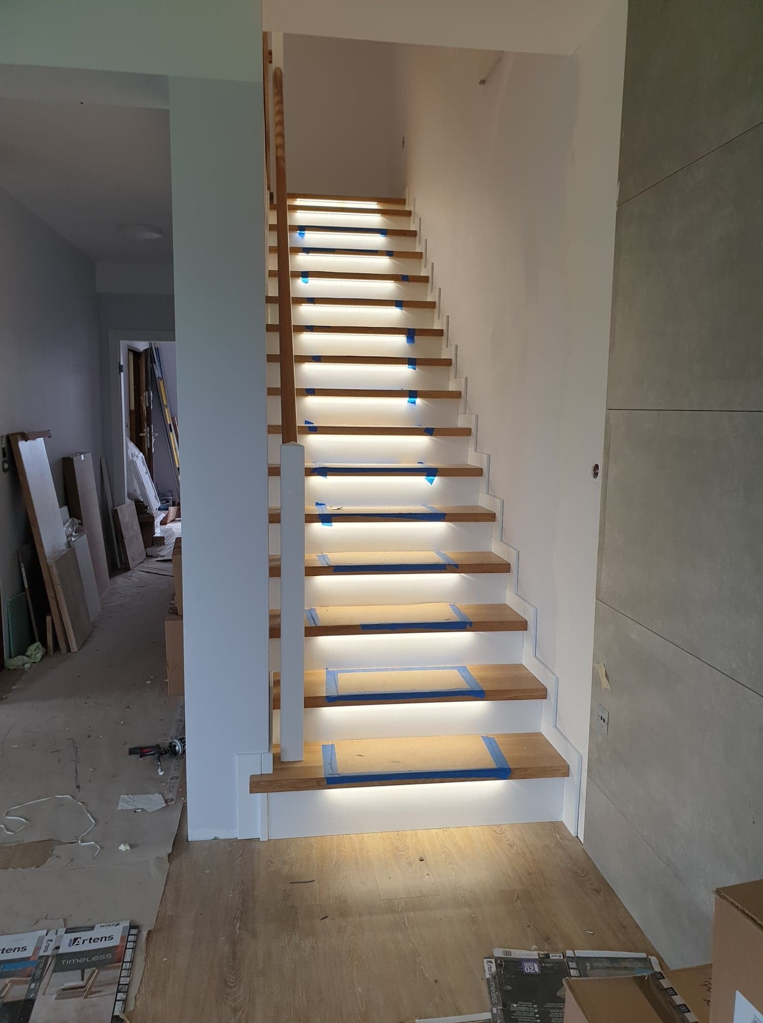 Drewniane schody z podświetleniem LED na każdym stopniu, zabezpieczone niebieską taśmą malarską, w trakcie montażu, widoczne narzędzia i materiały budowlane w tle.