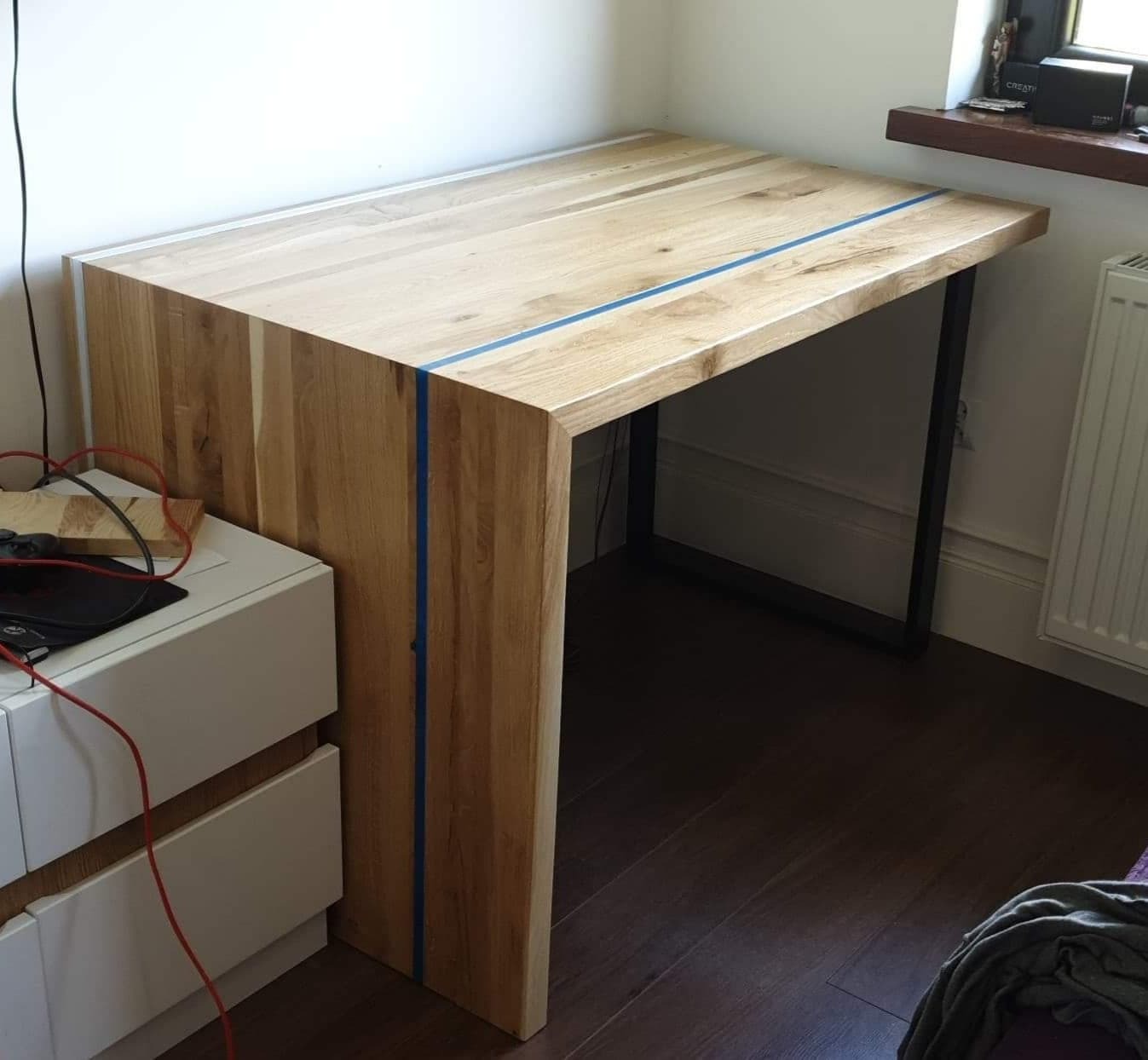 Drewniane biurko na metalowych nogach z niebieskim akcentem, stojące obok białej komody w pokoju z brązową podłogą.