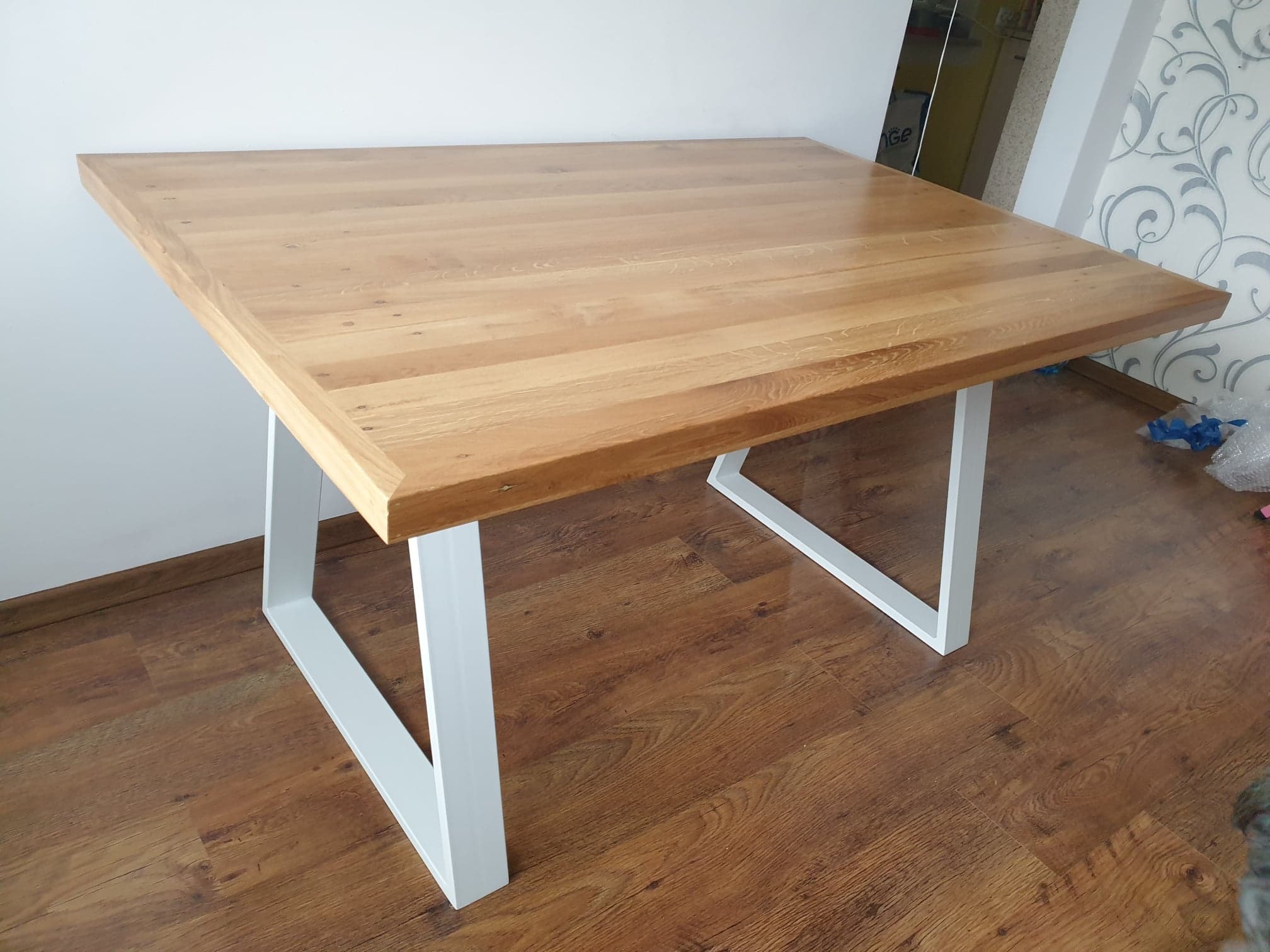 Kwadratowy stół z jasnym blatem drewnianym i białymi, metalowymi nogami w kształcie litery V, ustawiony na podłodze imitującej drewno.