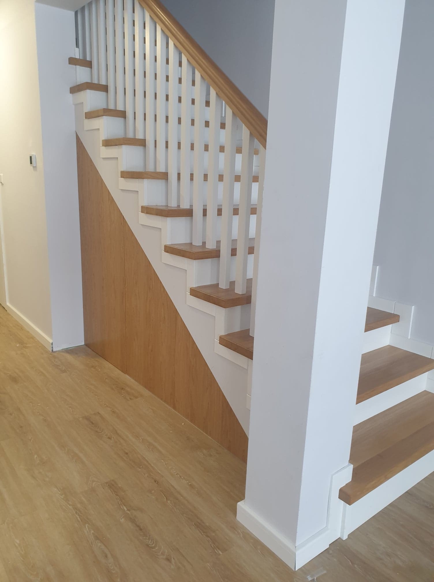 Nowoczesne schody drewniane z białymi balustradami i drewnianymi stopniami, z zabudową pod schodami w kolorze jasnego drewna, widok z dołu.
