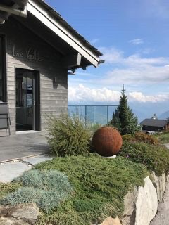 Nowoczesny dom z szarej deski z widokiem na góry, ozdobny ogród z kulą z rdzy i kamiennym murkiem oporowym.