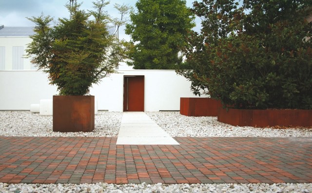 Nowoczesny ogród z geometrycznymi rabatami w kolorze rdzy, białym żwirem i ścieżką z jasnych płyt, prowadzącą do czerwonych drzwi w białym murze.