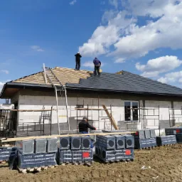 Budowa domu jednorodzinnego: murarz układa dachówkę na drewnianej konstrukcji dachu, na ziemi palety z dachówkami CREATON, ściany z betonu komórkowego, rusztowania.