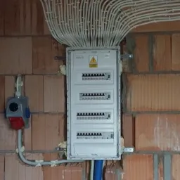 Rozdzielnica elektryczna z licznymi przewodami w peszlach, zamontowana na surowej ścianie z cegły, widoczna puszka instalacyjna z boku.