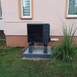 Passive Instal Sp. z o.o.- Mateusz Sztacherski - Czarna jednostka zewnętrzna pompy ciepła Panasonic Aquarea zainstalowana na betonowych blokach i żwirowym fundamencie przed beżową ścianą domu z dwoma oknami.