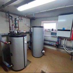 Passive Instal Sp. z o.o.- Mateusz Sztacherski - Dwa szare zbiorniki akumulacyjne ciepłej wody w kotłowni z ceramiczną podłogą, widoczne rury, zawory, manometry, oraz wiszący na ścianie biały piec grzewczy z niebieskimi akcentami.