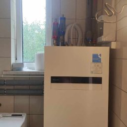 Passive Instal Sp. z o.o.- Mateusz Sztacherski - Biała pompa ciepła Panasonic w pomieszczeniu z ceramicznymi płytkami, widoczne rury i filtry, obok pralka
