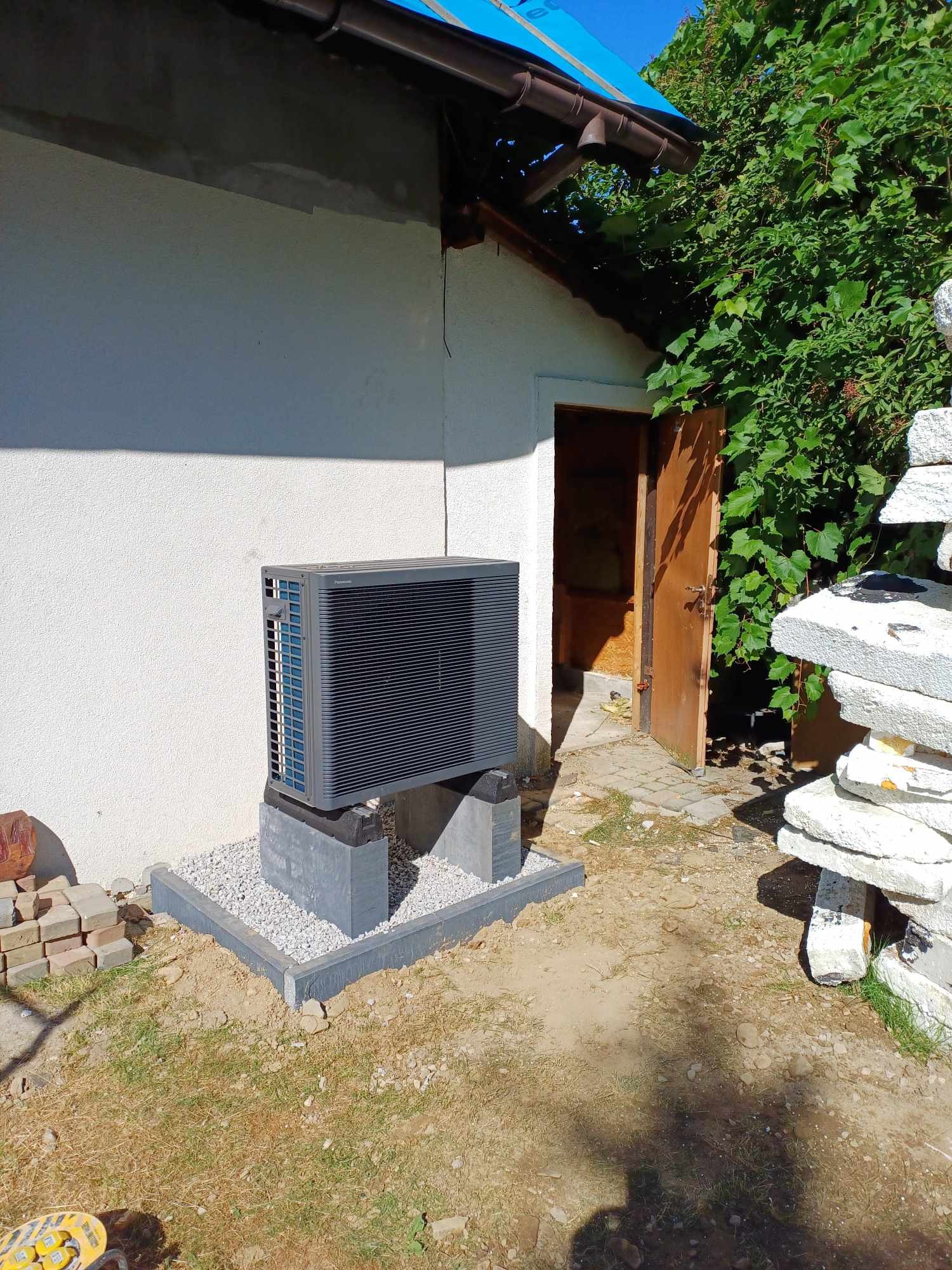 Zewnętrzna jednostka klimatyzacji/pompy ciepła marki Panasonic, zainstalowana na betonowym cokole z obramowaniem wypełnionym białym żwirem, przy białej ścianie budynku z otwartymi drzwiami i stertą...