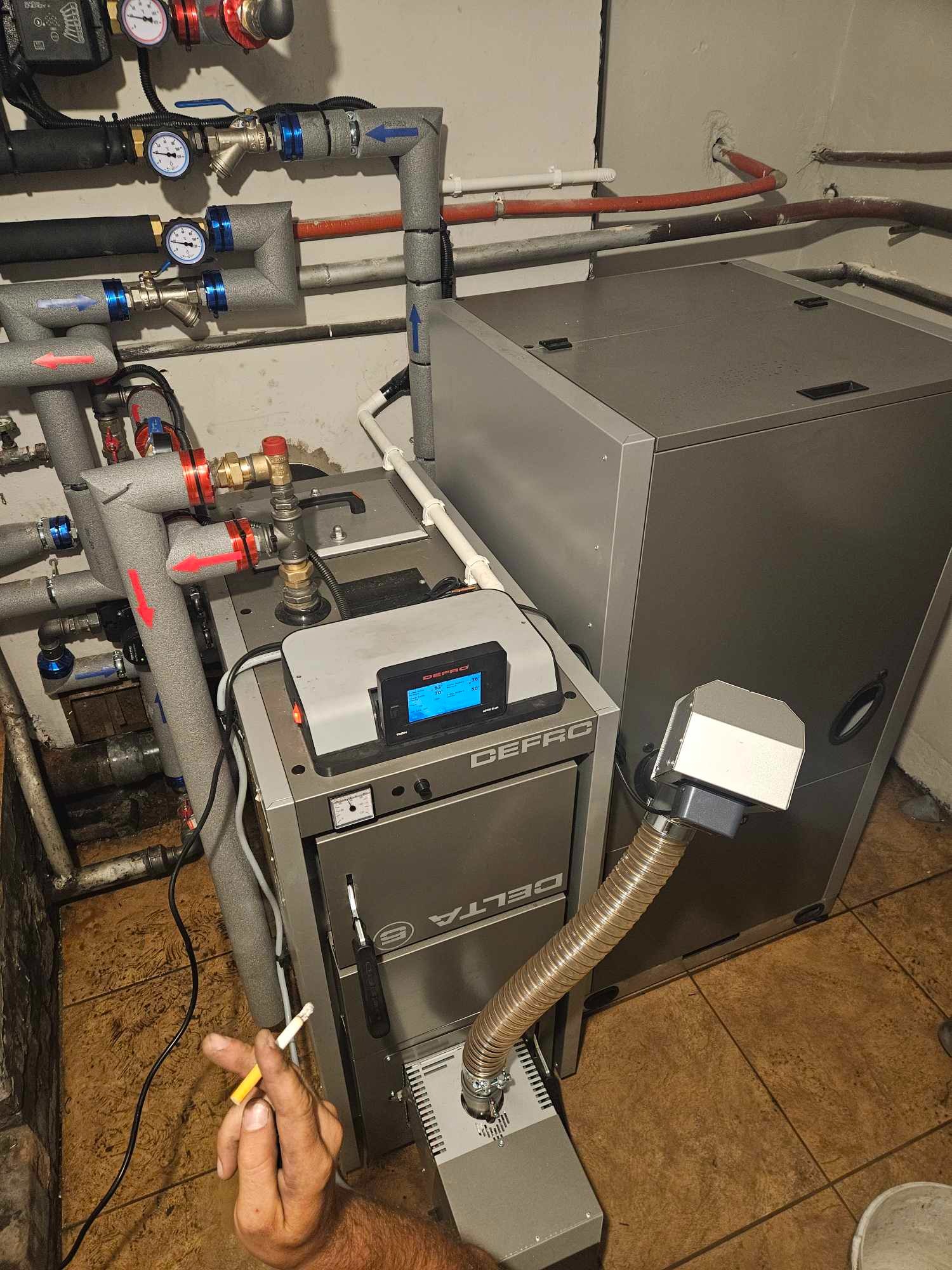 Szary piec Defro Delta S z podłączonymi rurami, widoczne manometry i ekran sterujący, w tle drugi piec, w kadrze dłoń trzymająca papierosa.