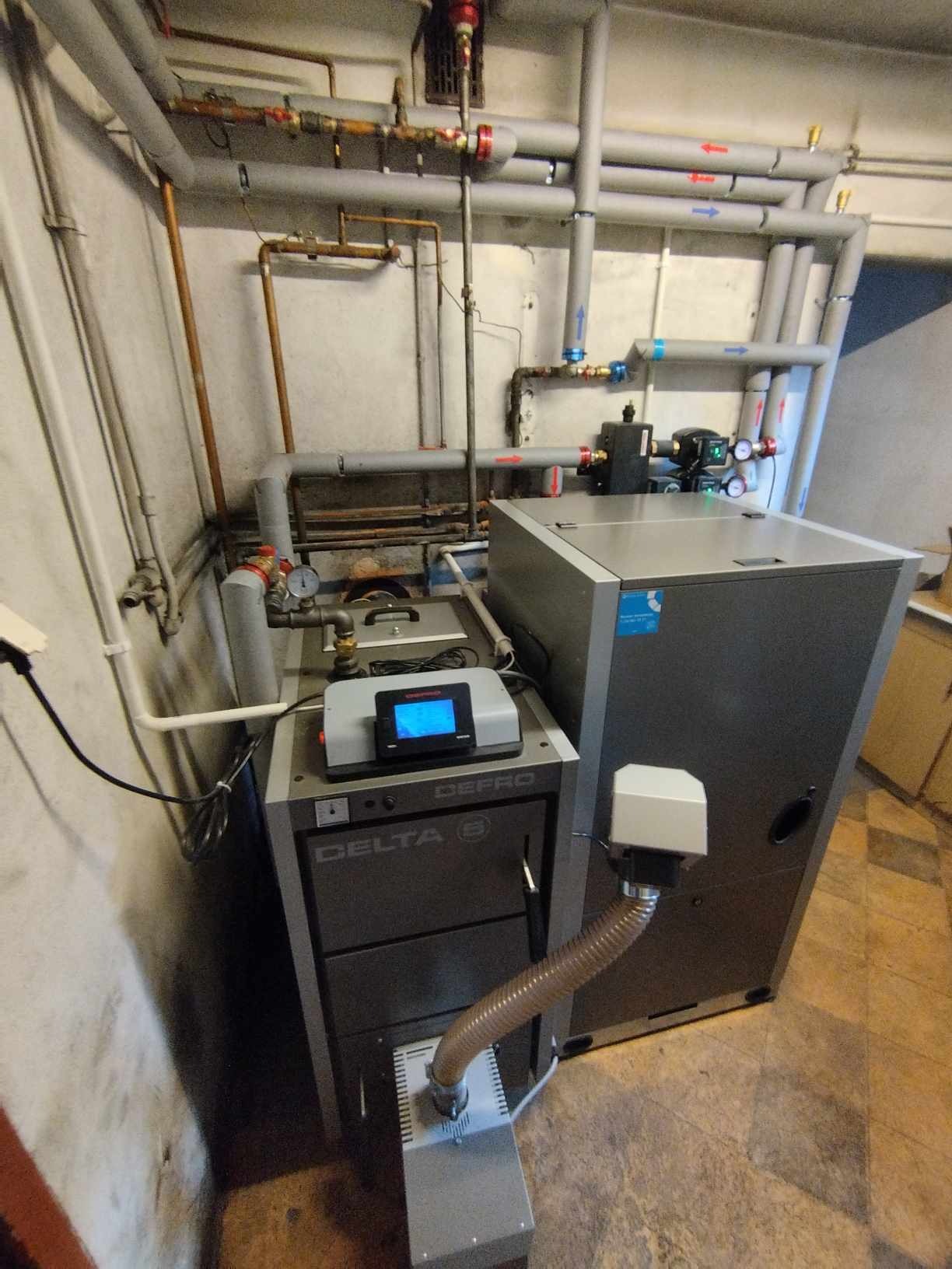 Szara kotłownia z dwoma piecami Defro Delta 8, licznymi rurami grzewczymi i przewodami, widoczne manometry i zawory.