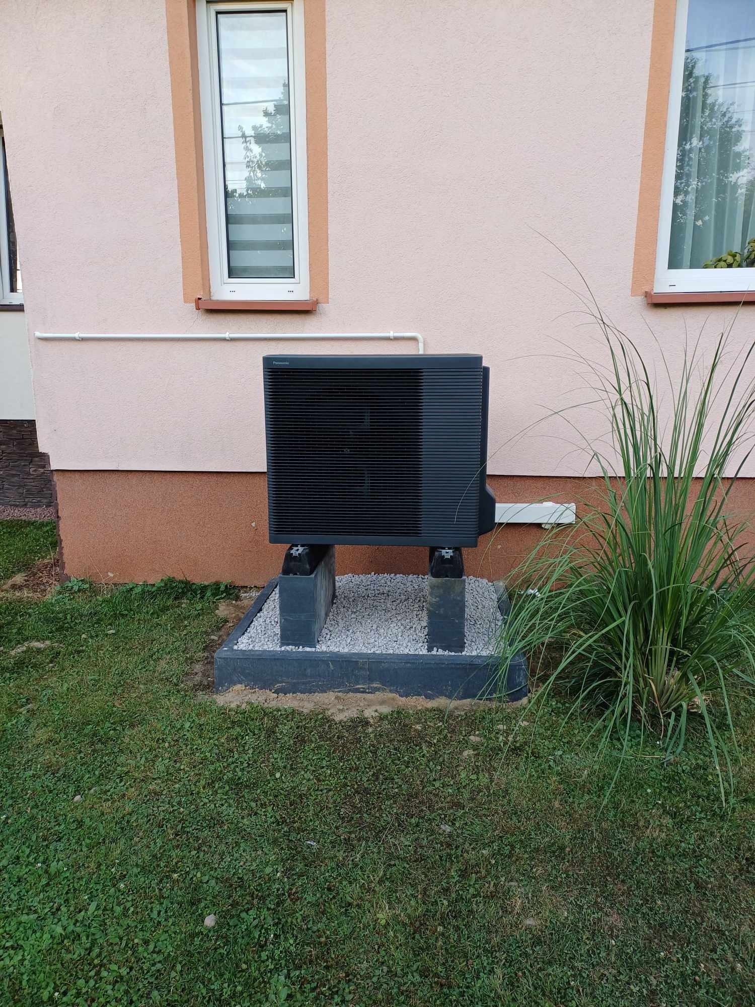 Czarna jednostka zewnętrzna pompy ciepła Panasonic Aquarea zainstalowana na betonowych blokach i żwirowym fundamencie przed beżową ścianą domu z dwoma oknami.