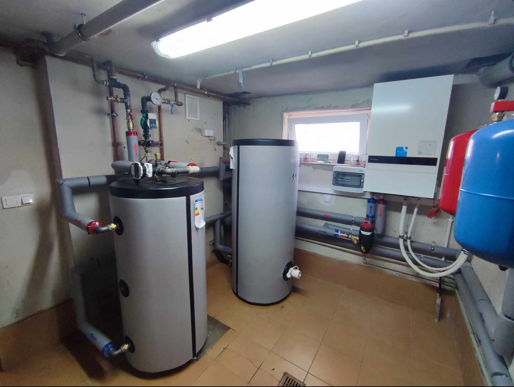 Dwa szare zbiorniki akumulacyjne ciepłej wody w kotłowni z ceramiczną podłogą, widoczne rury, zawory, manometry, oraz wiszący na ścianie biały piec grzewczy z niebieskimi akcentami.