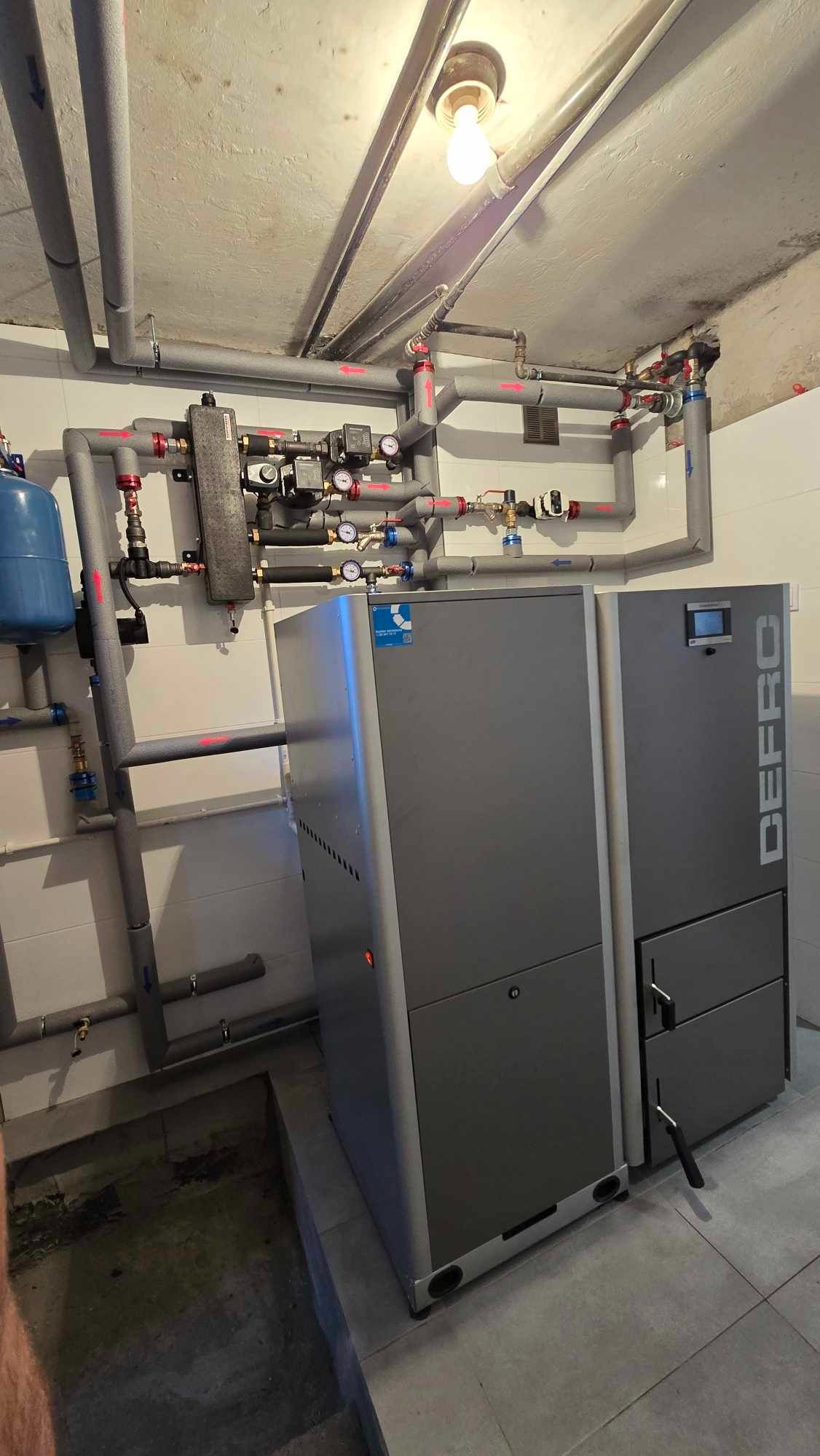 Szary piec CO marki DEFRO z podajnikiem, zainstalowany w kotłowni z rurami grzewczymi i armaturą, oświetlony żarówką na suficie.