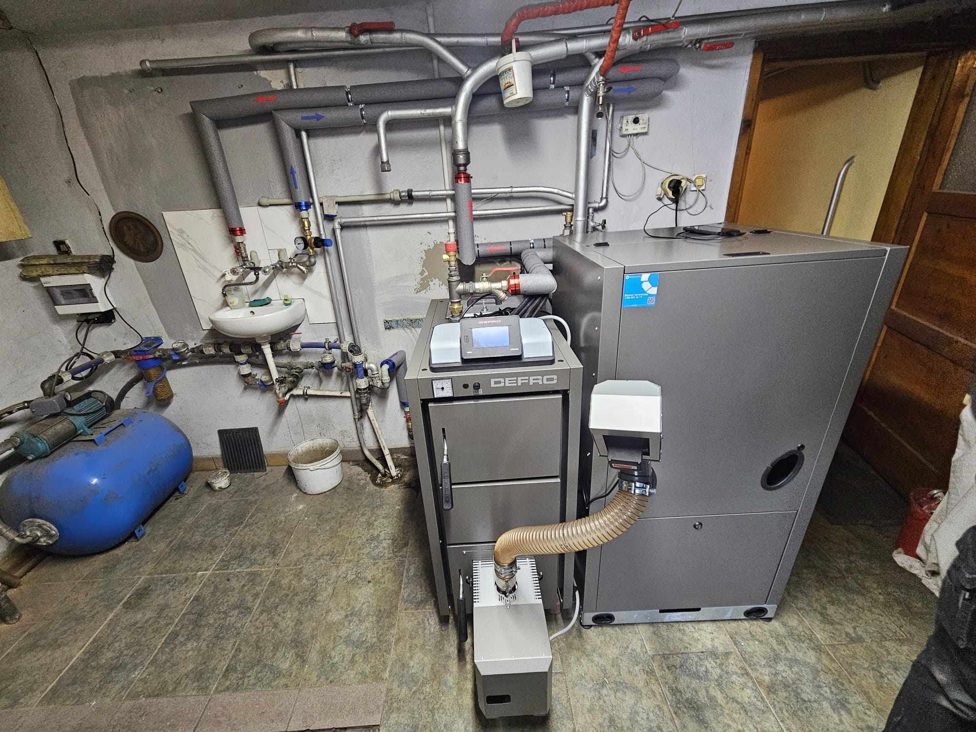 Nowoczesny piec DEFRO z podajnikiem ślimakowym w kotłowni, widoczne rury instalacji grzewczej, umywalka i niebieski zbiornik ciśnieniowy, z boku fragment otwartych drzwi.