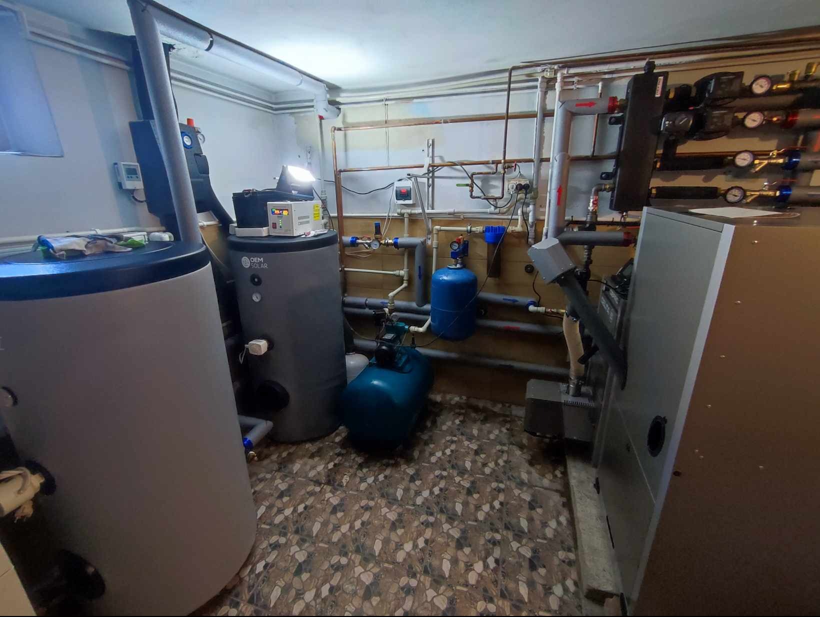 Zestaw zbiorników akumulacyjnych w kotłowni z licznymi rurami, zaworami i manometrami, z widocznym logo OEM Solar na jednym ze zbiorników.