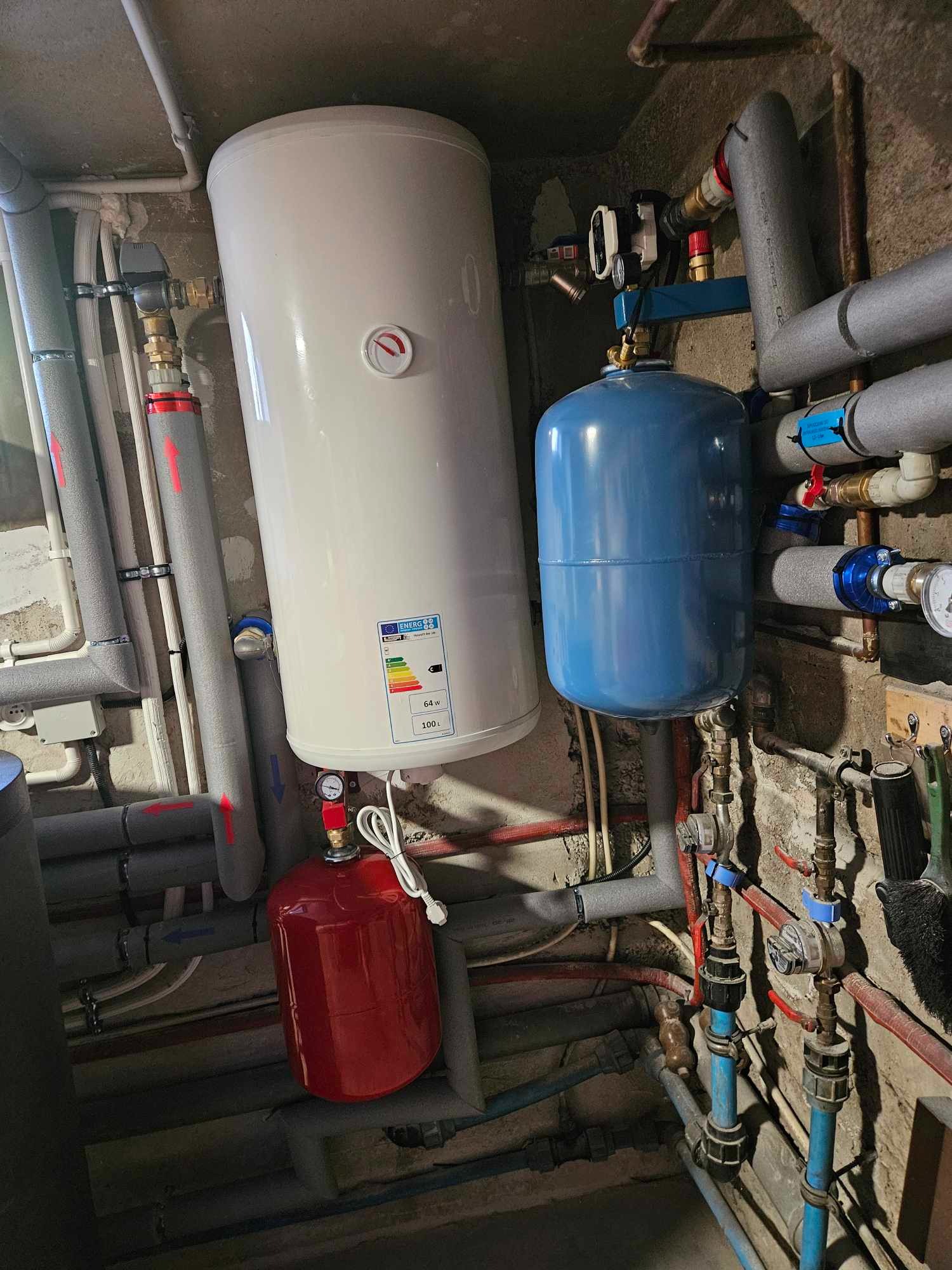 Biały bojler z etykietą energetyczną, czerwony i niebieski zbiornik wyrównawczy w kotłowni z licznymi rurami.