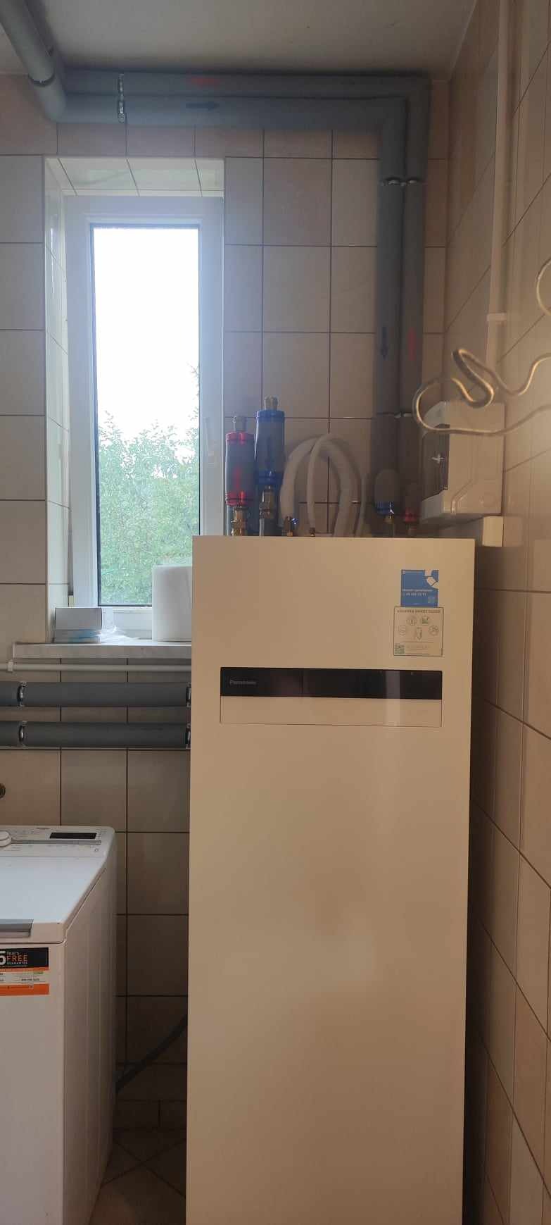 Biała pompa ciepła Panasonic w pomieszczeniu z ceramicznymi płytkami, widoczne rury i filtry, obok pralka
