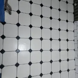 Białe, kwadratowe płytki podłogowe z czarnymi, romboidalnymi wstawkami w łazience, widoczne rury i fragment niebieskiej tkaniny w lewym górnym rogu, ujęcie z góry.