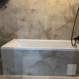 Biała wanna obudowana płytkami imitującymi marmur, z czarną armaturą. Na ścianie widoczny piec gazowy. Spokojna kolorystyka i eleganckie wykończenie łazienki.