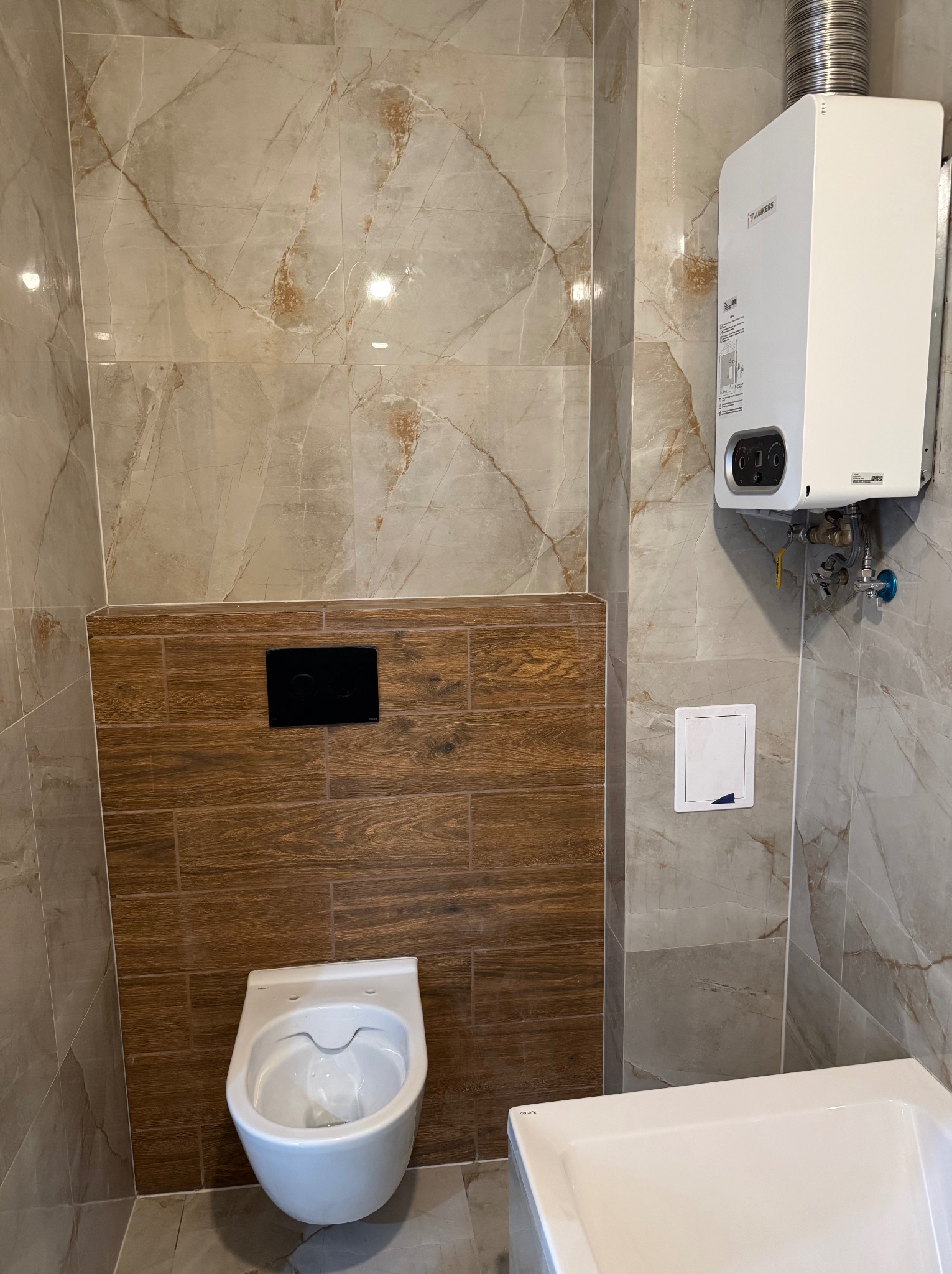 Wykończona łazienka z beżowymi płytkami imitującymi marmur i drewnopodobnymi panelami za wiszącą toaletą. Widoczny bojler i fragment wanny.