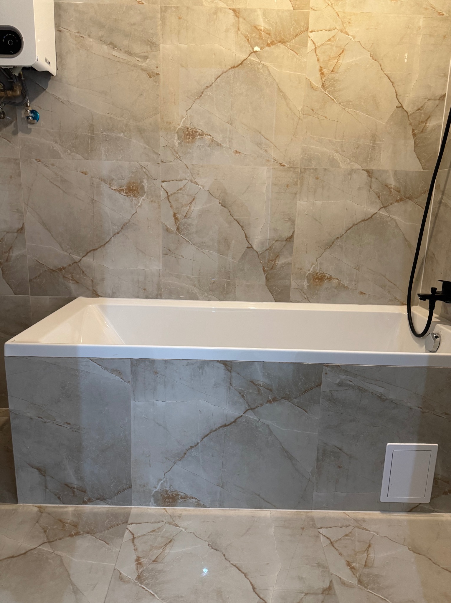 Biała wanna obudowana płytkami imitującymi marmur, z czarną armaturą. Na ścianie widoczny piec gazowy. Spokojna kolorystyka i eleganckie wykończenie łazienki.