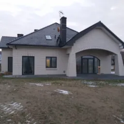 Nowoczesny dom jednorodzinny z białej cegły klinkierowej, czarnym dachem z dachówki i podcieniem wejściowym z łukiem. Na dachu widoczna antena satelitarna. Teren wokół domu częściowo pokryty śniegiem.