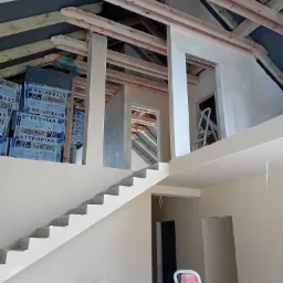 Wnętrze domu w trakcie prac wykończeniowych: ściany po nałożeniu jasnego tynku, widoczne drewniane elementy konstrukcji dachu, schody w surowym stanie i izolacja styropianowa w niebieskich...