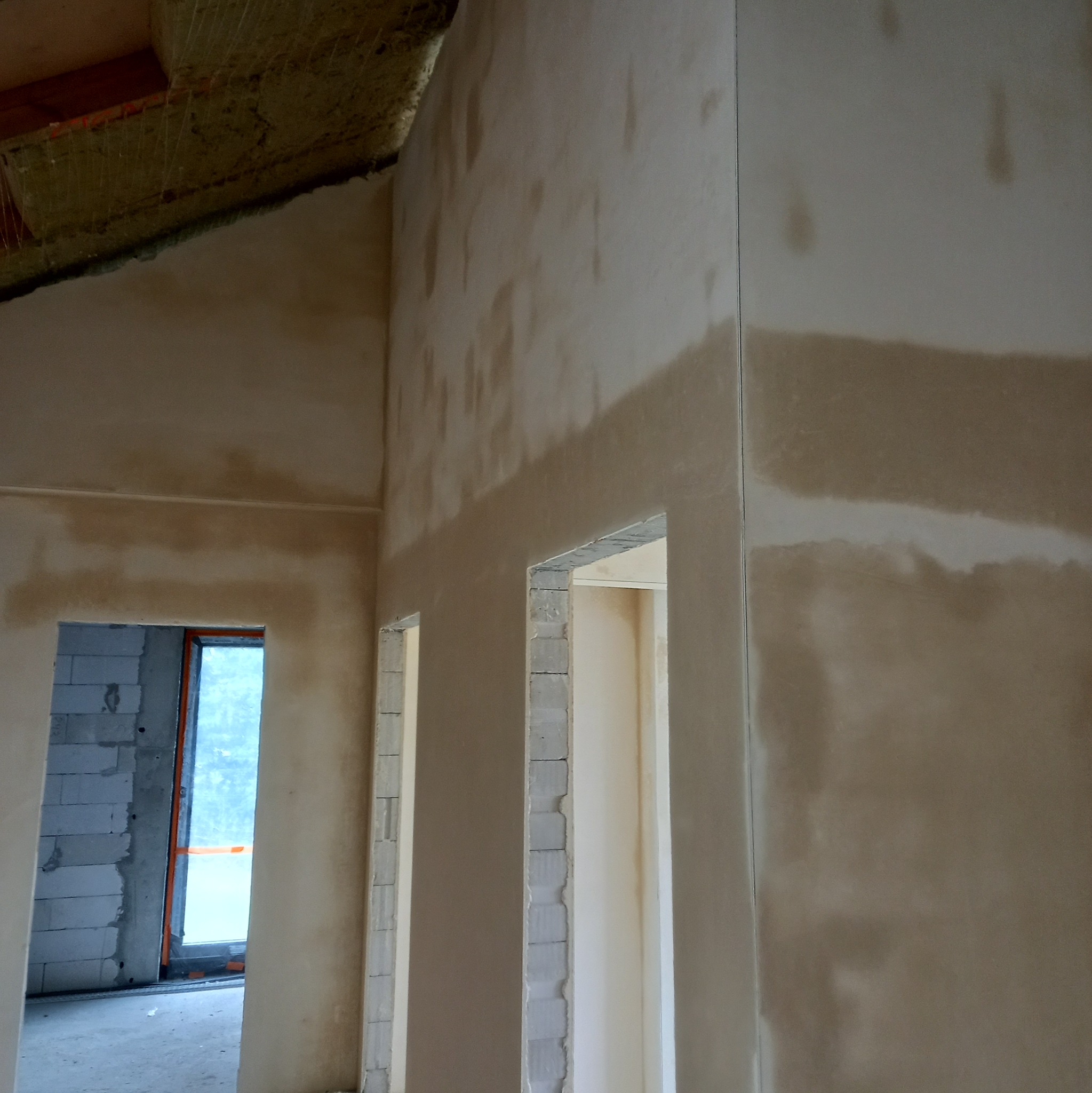 Ściany w trakcie prac wykończeniowych, widoczne ślady po nałożonym tynku, surowy stan budynku z widocznymi otworami drzwiowymi i oknem w głębi.