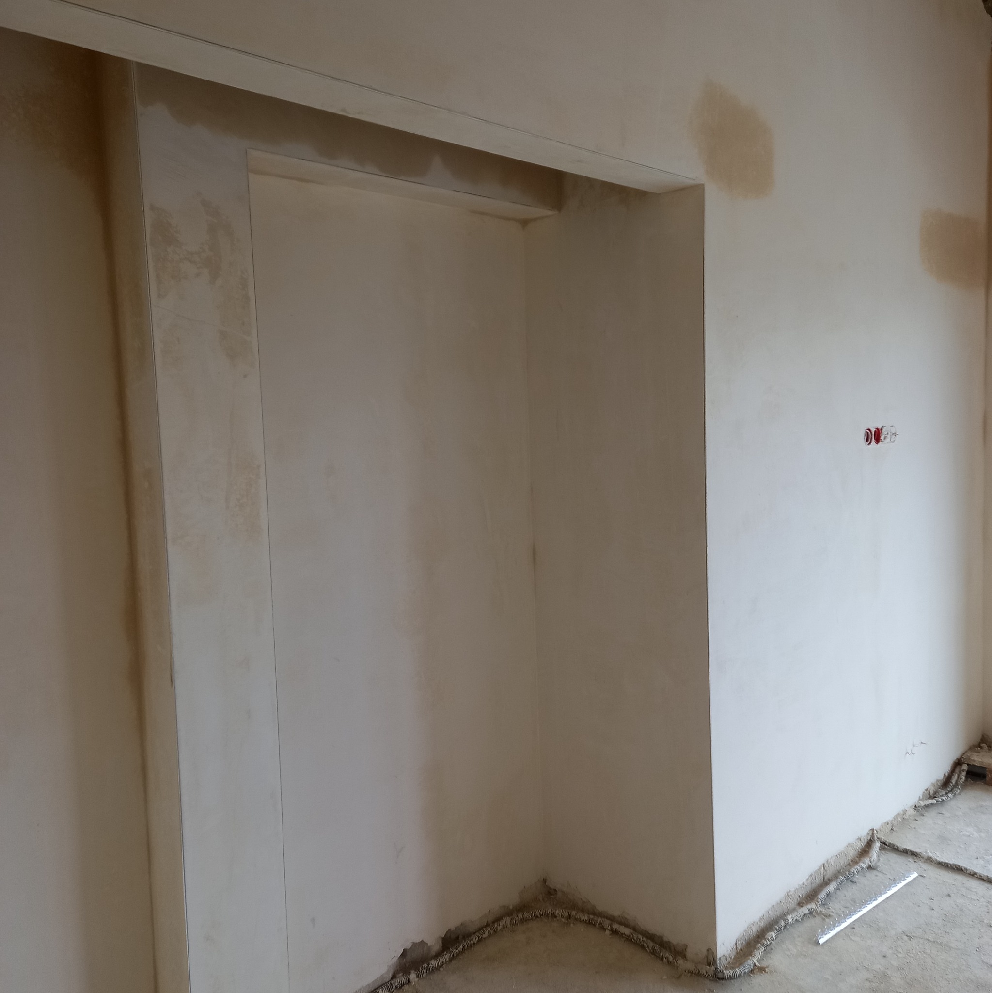 Świeżo nałożony tynk maszynowy na ścianach i ościeżnicy wewnątrz remontowanego pomieszczenia, widoczne ślady po łatach i gniazdka elektryczne bez osprzętu.