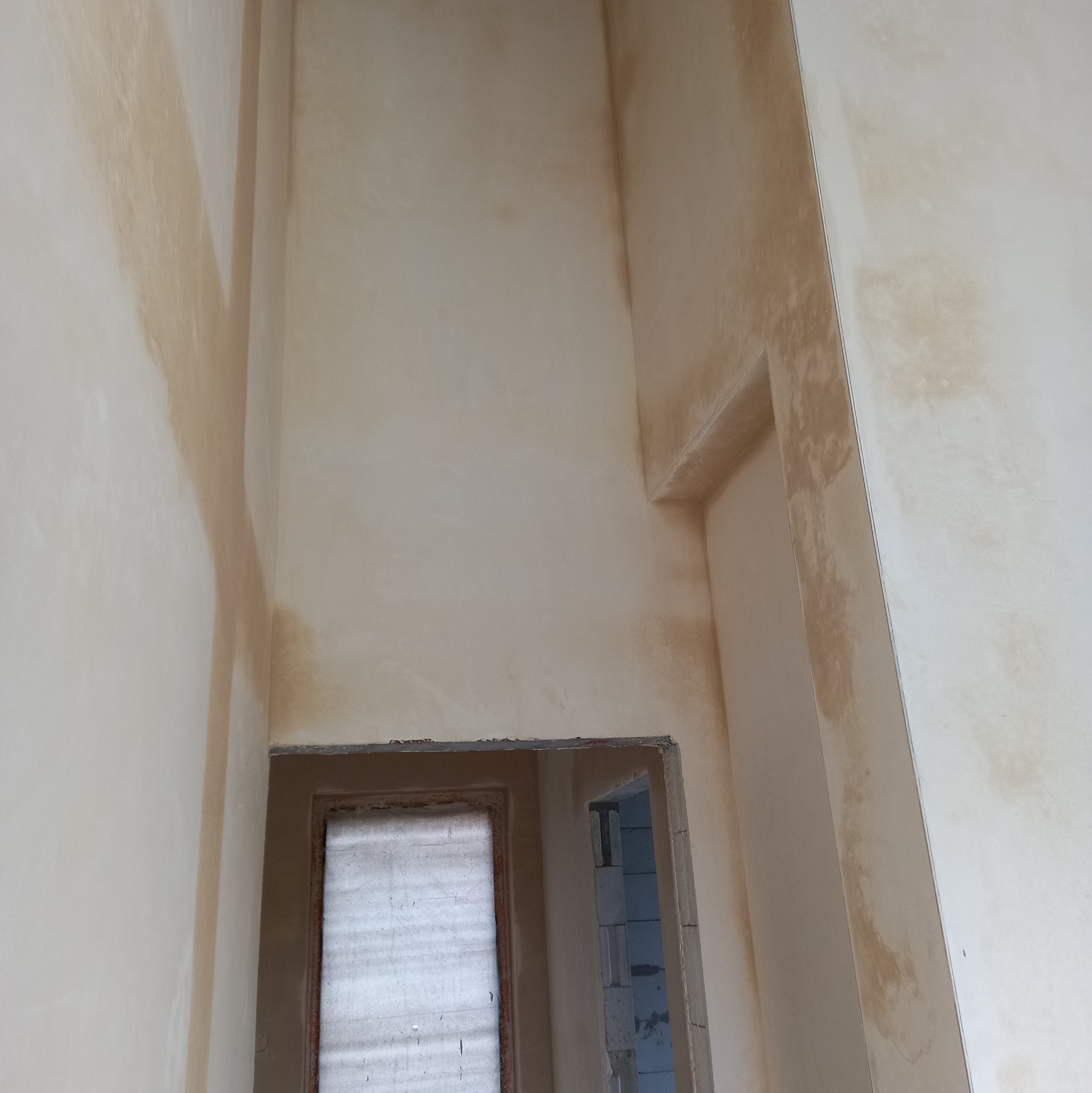 Świeżo nałożone, wilgotne tynki maszynowe w jasnym odcieniu, widoczne ślady wilgoci i nierównomierne wysychanie na ścianach i wokół ościeżnic okiennych i drzwiowych.