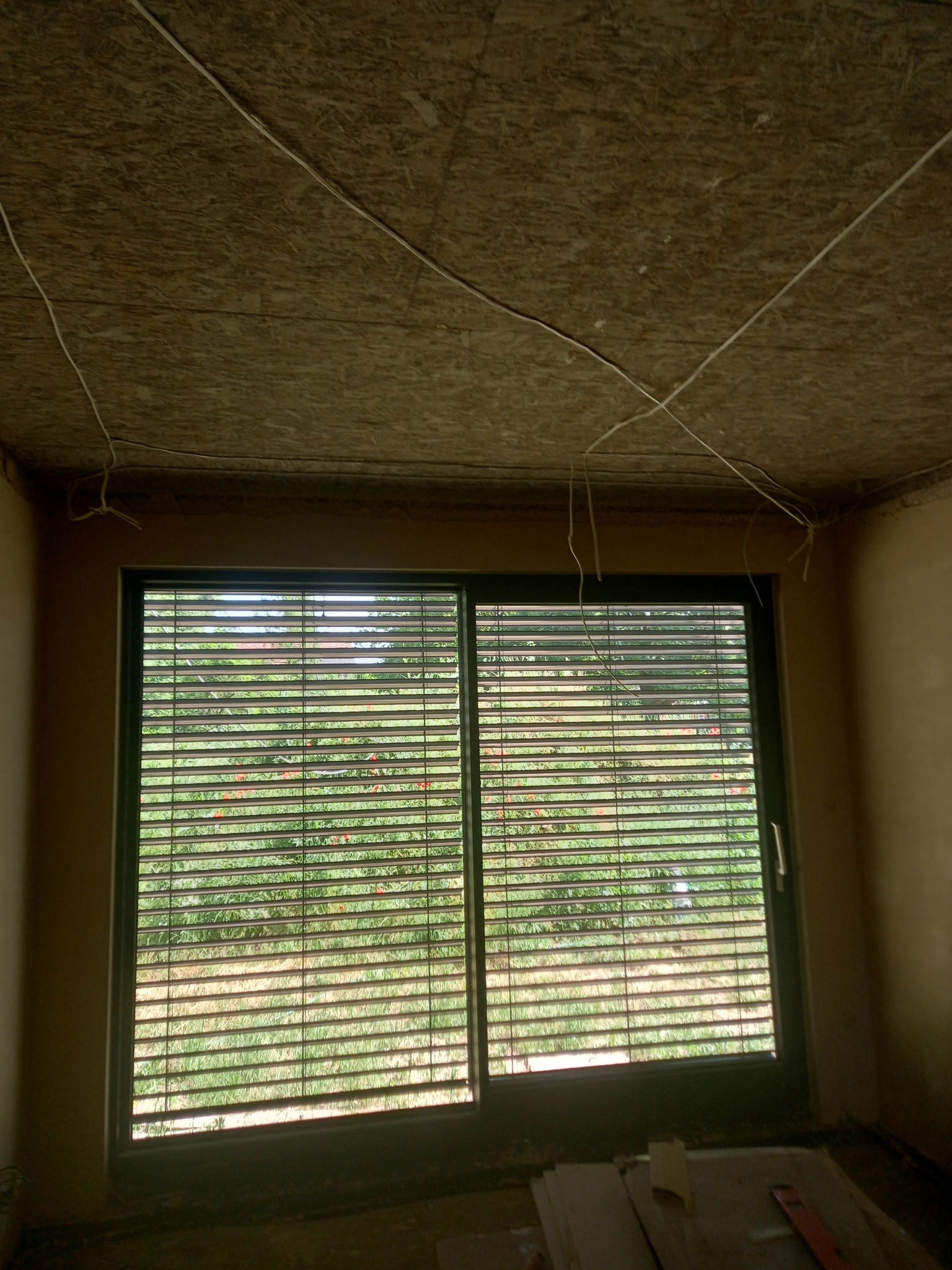 Surowy strop z płyty OSB z wystającymi kablami elektrycznymi nad oknem z żaluzjami, w pokoju przed wykończeniem ścian.