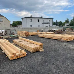 Składowane drewno budowlane na placu, widoczne ułożone stosy tarcicy i pnie drzew, w tle budynki mieszkalne i fragment ogrodzenia.