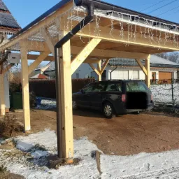 Drewniana wiata garażowa z oświetleniem świątecznym, częściowo pokryta śniegiem, z zaparkowanym ciemnym kombi.