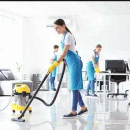 Zespół sprzątający w jasnym biurze: kobieta odkurza błyszczącą podłogę, a w tle dwie osoby przecierają biurka, wszyscy w niebieskich fartuchach.